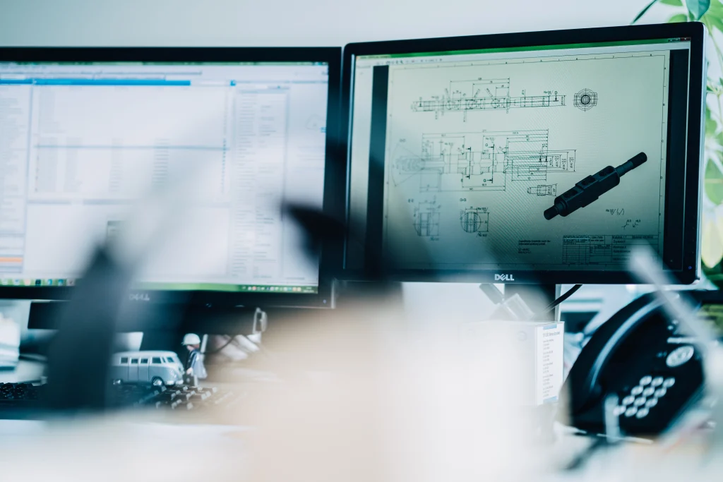 Zwei Computermonitore auf einem Büroschreibtisch, einer zeigt eine technische CAD-Zeichnung eines Maschinenteils, der andere eine Tabellenkalkulation