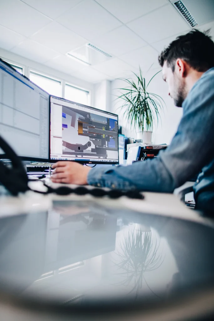 Mitarbeiter am Schreibtisch arbeitet an einem CAD-Programm mit 3D-Maschinenmodell auf dem Monitor in einem hellen Büro