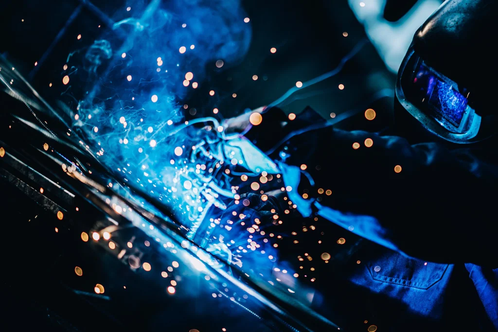 Schweißer mit Schutzhelm beim Schweißen von Stahl mit blauem Lichtbogen und Funkenflug vor dunklem Hintergrund